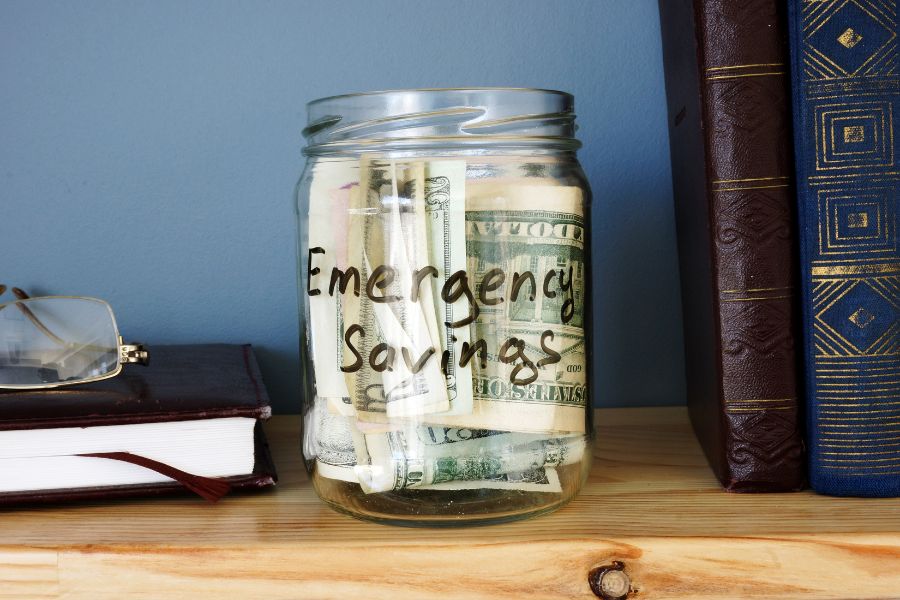 7 Easy Ways to Save Money at the Vet and the Expensive Mistake Almost Every Pet Owner Is Making 6 A labeled jar filled with cash sits on a wooden shelf beside books and glasses. Building an emergency savings fund is one of the most effective ways to save money at the vet when unexpected costs arise.